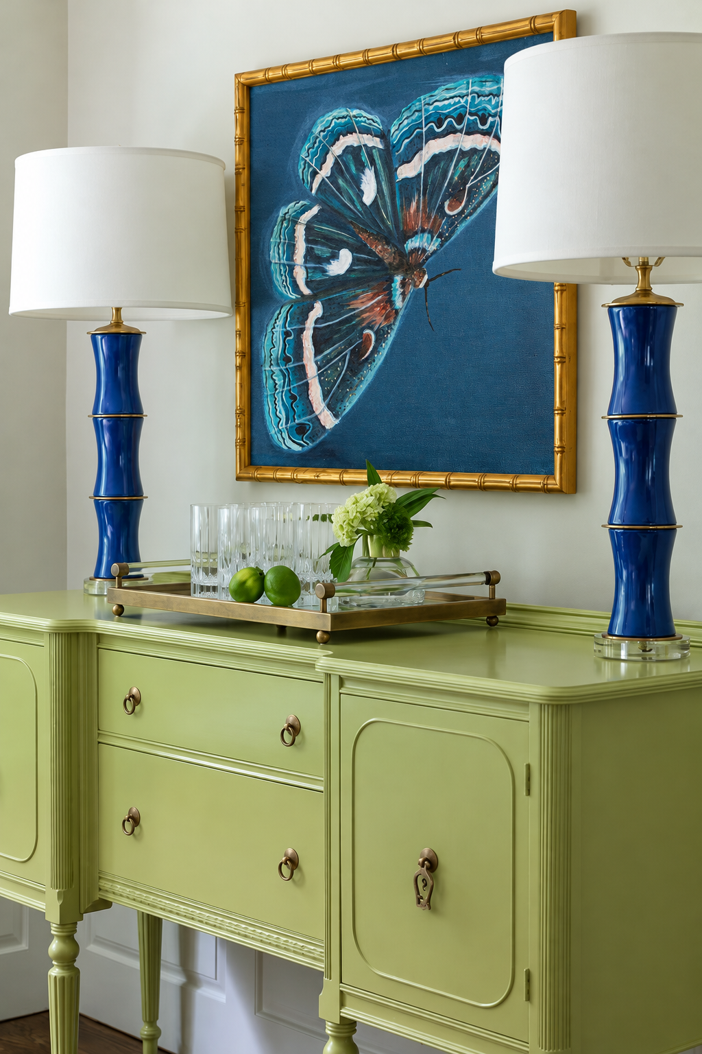 Justice blue moth artwork in a gold frame above a pale green sideboard between two cobalt blue lamps