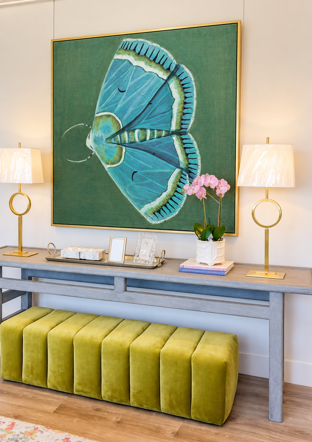 Prudence blue-green moth canvas in a gold floating frame above an elegant console with ivory lamps and pink orchids